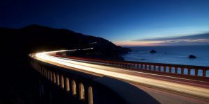 long exposure photo of a bridge over the ocean with head lights extending the entire length of the road
