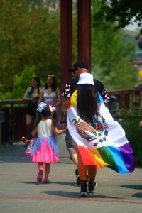 a person with long hair with a queer pride mexican flag holds hands with a child facing away from the camera