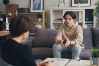 A teenager is sitting on a couch in a comfortable therapeutic space talking with a counsellor who is holding a clipboard.